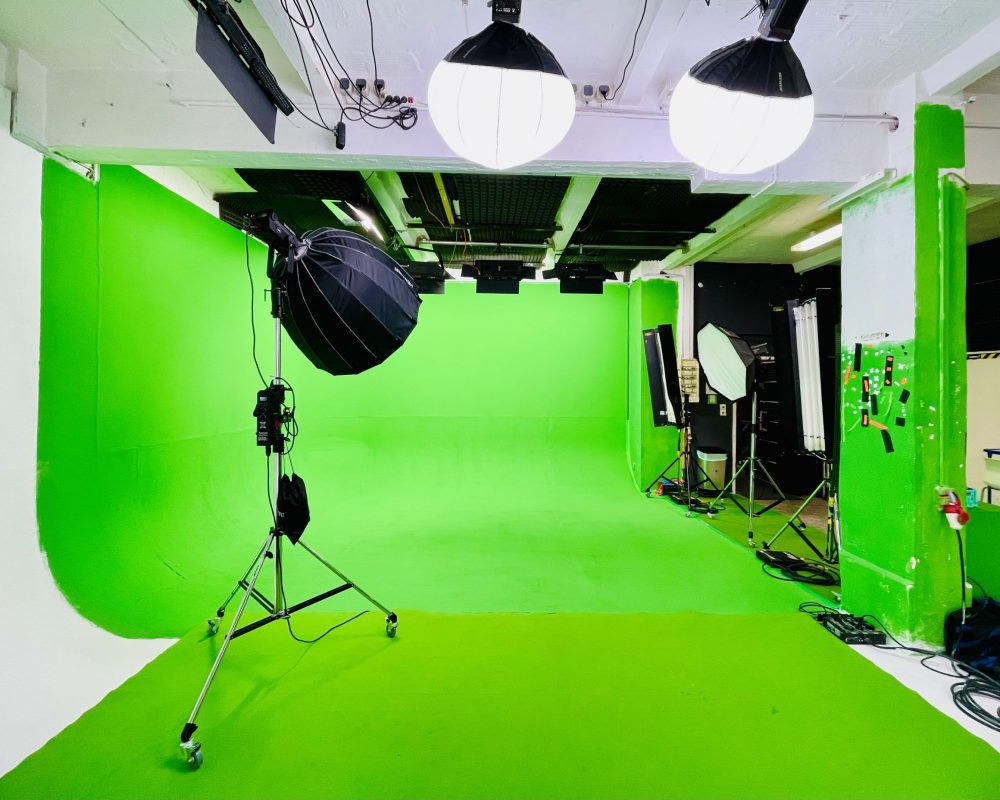 Grüne Eckhohlkehle im Greenscreen-Studio, der Hintergrund ist gleichmäßig ausgeleuchtet, es hängen zwei leuchtende Studiolampen mit Laternen-Softbox an der Decke, auf der linken Seite steht eine Aputure 300x mit einer Parabolic Octabox bereit, rechts vom Greenscreen weitere Lichttechnik. Ansicht aus der weißen Ecke der Hohlkehle heraus